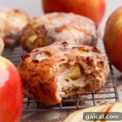 A close-up of a perfectly glazed apple pastry, reminiscent of a gourmet air fryer apple fritter, with a tantalizing bite taken out. It rests elegantly on a cooling rack, artfully surrounded by fresh whole apples and thinly sliced apple segments, beautifully evoking warm, comforting autumn flavors.