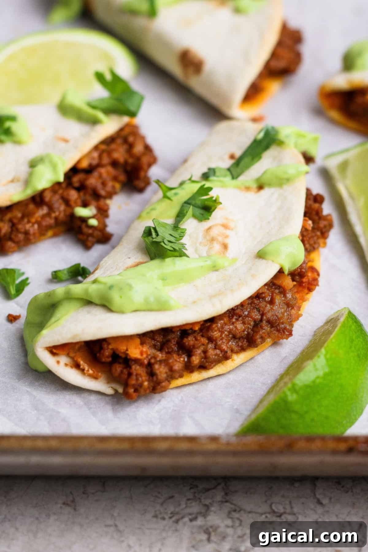 A crispy sheet pan taco filled with seasoned ground meat, topped with vibrant green avocado crema and fresh cilantro, served on a baking sheet with lime wedges, highlighting a perfect weeknight meal.