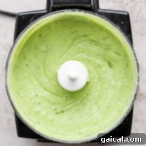 Top view of a food processor containing smooth, creamy green sauce or dip, likely a blended avocado or herb mixture.