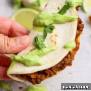 A hand holds a taco filled with ground beef and topped with green sauce and chopped cilantro on a light surface.