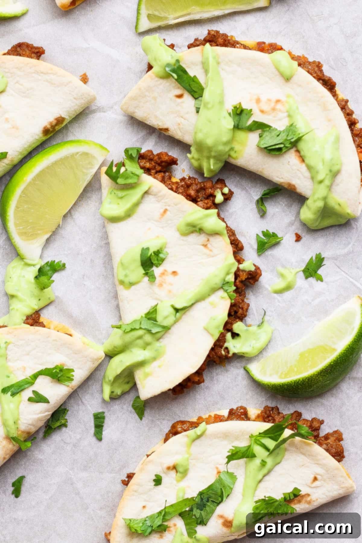 Three perfectly folded and baked sheet pan tacos filled with seasoned ground beef, garnished with vibrant green avocado crema and fresh cilantro, neatly arranged on parchment paper with bright lime wedges.