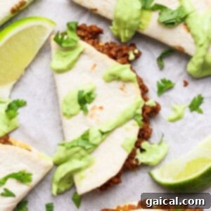 Taco halves with ground meat filling, topped with green sauce and cilantro, served with lime wedges on a white surface.