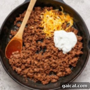 Ground beef in a cast iron skillet with shredded cheese and a dollop of sour cream, being stirred with a wooden spoon.