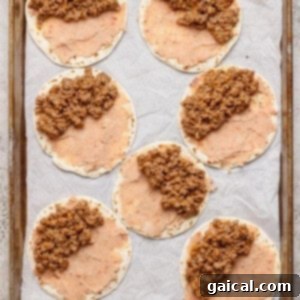 Seven tortillas spread with refried beans and ground beef mixture are arranged on a parchment-lined baking sheet.