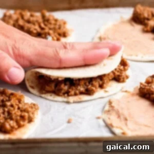A hand folds a tortilla filled with ground meat mixture on a baking sheet lined with parchment paper, with more tortillas being assembled nearby.