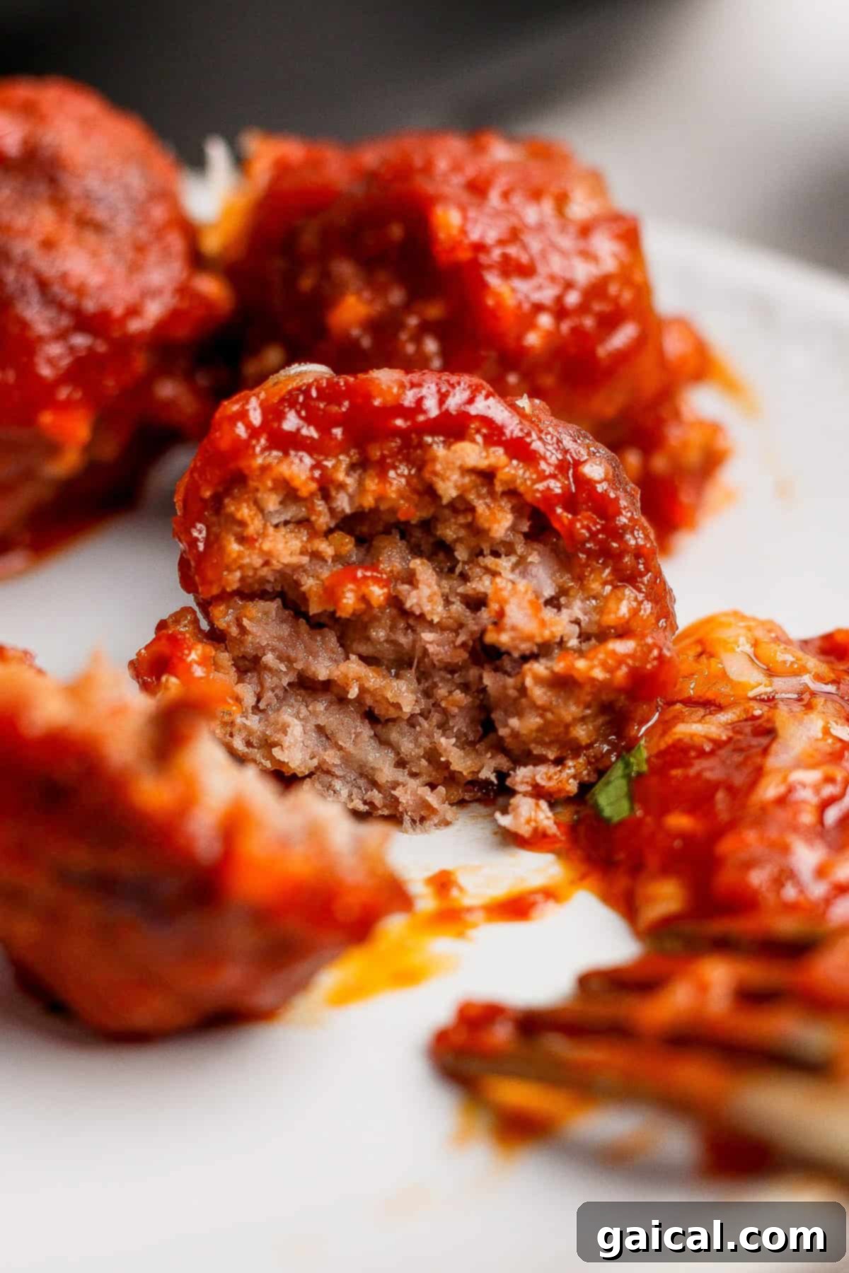 A tantalizing close-up of a serving of sloppy joe meatballs coated in rich, glossy tomato sauce on a white plate. One meatball is cut open, revealing its perfectly cooked, tender interior and inviting texture.