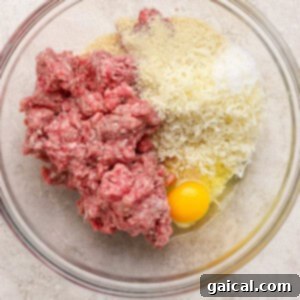 A glass bowl containing raw ground beef mixture for meatballs, with grated cheese, breadcrumbs, and a whole egg clearly visible on a light-colored surface, ready for mixing.