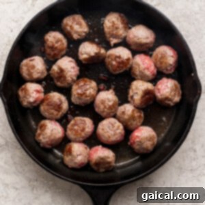 A black cast iron skillet contains several beef meatballs in the process of cooking. Some meatballs are already browned on one side, while others still show their raw, pinkish hue, indicating they are being turned.