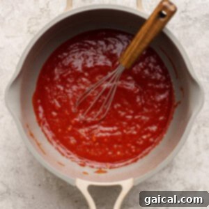 A silver saucepan containing a vibrant red tomato sauce mixture, with a metal whisk resting inside, indicates the sauce is being prepared on a light-colored kitchen surface.