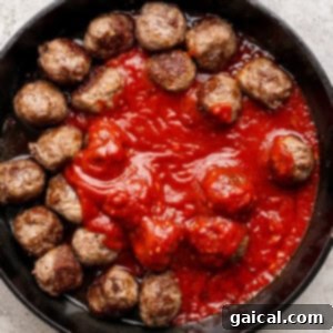 A cast iron skillet holds perfectly browned meatballs artfully arranged around a pool of rich, red tomato sauce, suggesting they are about to be combined and simmered.