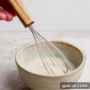 Gourmet Strawberry Toaster Tarts 14 A hand holding a whisk stirring a light-colored mixture in a ceramic bowl on a light surface.