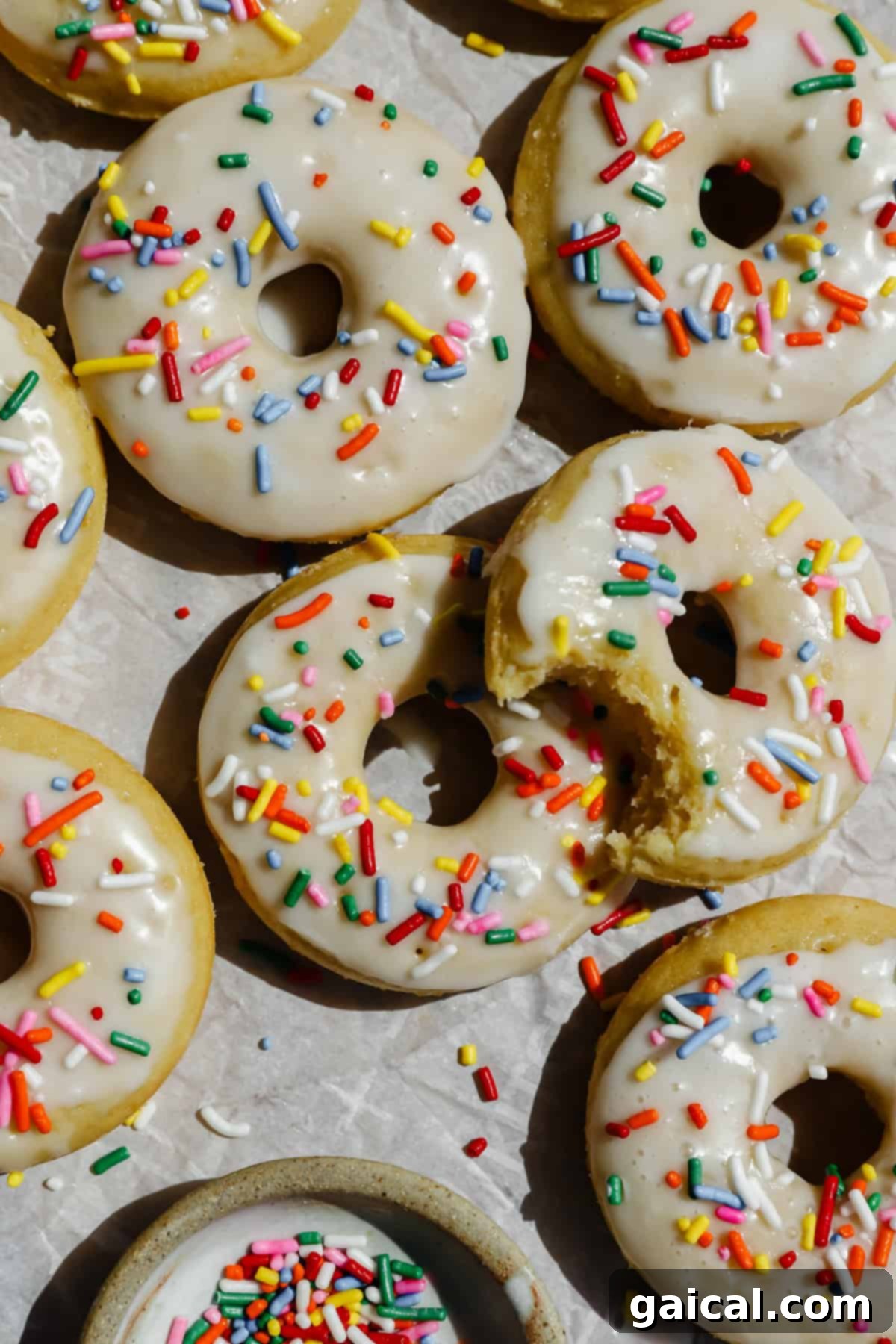 Glazed donuts topped with colorful sprinkles are arranged on parchment paper, with one donut partially eaten and a small bowl of extra sprinkles nearby. The donuts look soft and inviting.