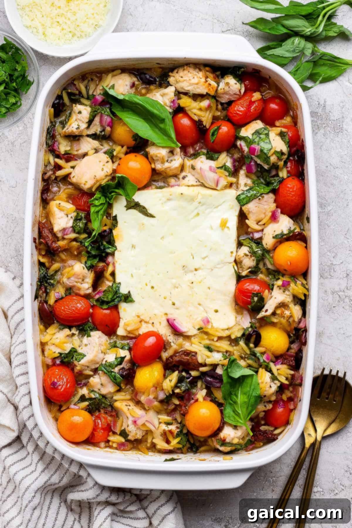 Zesty Greek Chicken and Orzo Bake 2 A baked casserole dish filled with tender chicken, vibrant cherry tomatoes, rich orzo pasta, crisp red onions, and a large block of creamy melted feta cheese in the center, garnished with fresh basil leaves.