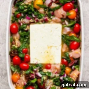 A white baking dish filled with raw chicken pieces, cherry tomatoes, spinach, chopped red onions, orzo, and a large block of feta cheese in the center.