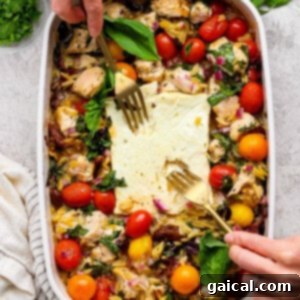 A person uses two forks to serve a baked dish with chicken, cherry tomatoes, greens, orzo, and feta cheese in a white casserole dish. Fresh herbs are visible on the side.