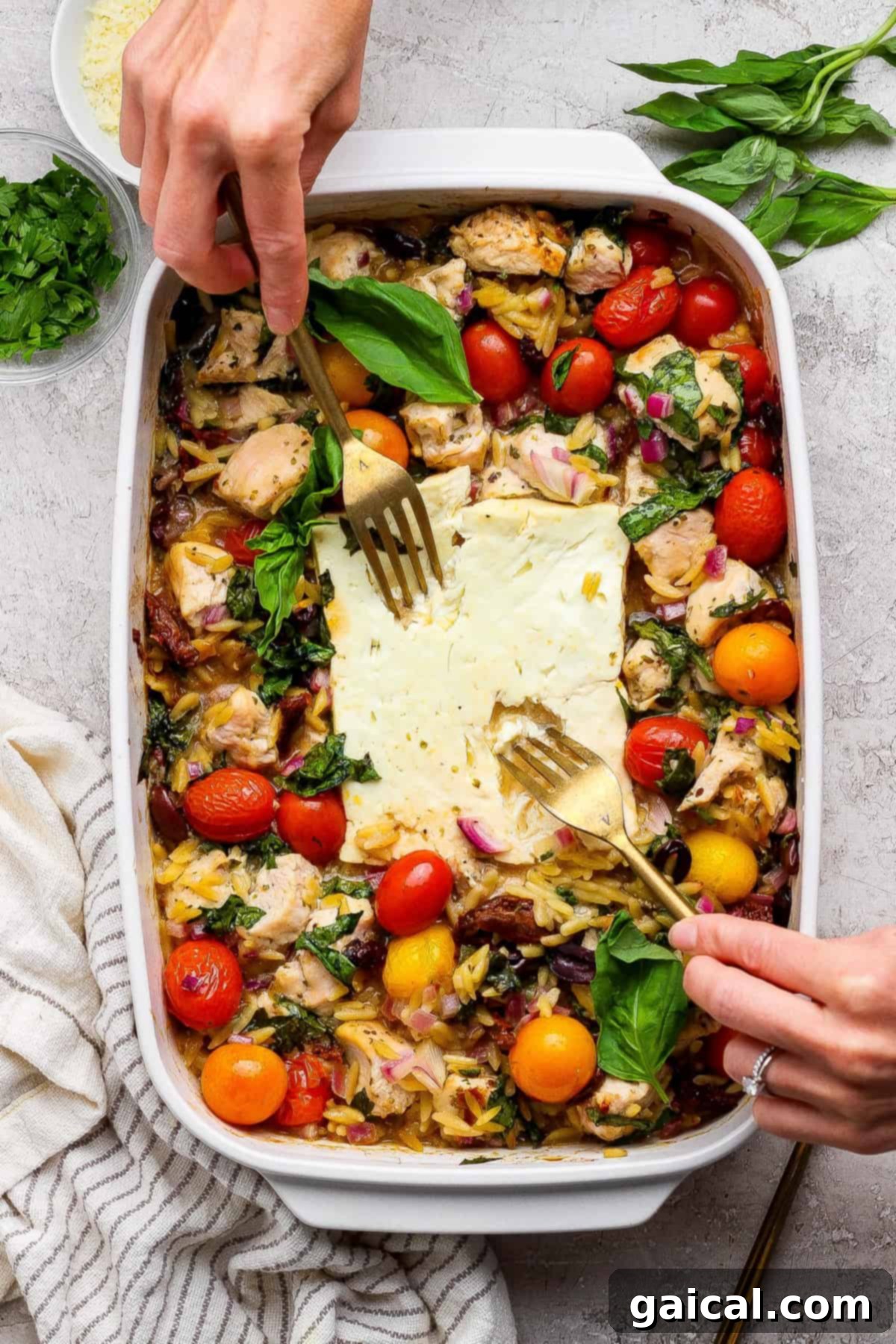 Zesty Greek Chicken and Orzo Bake 5 A person uses two forks to serve a baked dish with succulent chicken pieces, vibrant cherry tomatoes, fresh greens, perfectly cooked orzo, and rich feta cheese from a white casserole dish. Fresh herbs are visible on the side, highlighting the garnish.