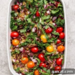 A white baking dish filled with fresh spinach, cherry tomatoes, chopped red onion, sun-dried tomatoes, and olives on a light textured surface.