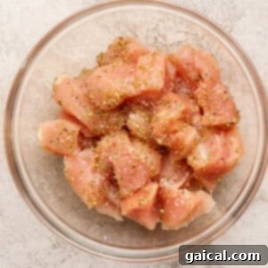 A glass bowl filled with raw, diced chicken breast pieces, seasoned with salt, pepper, and dried herbs, on a light textured surface.