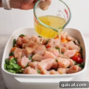 A hand pours broth from a glass measuring cup over raw chopped chicken, tomatoes, and greens in a white baking dish.