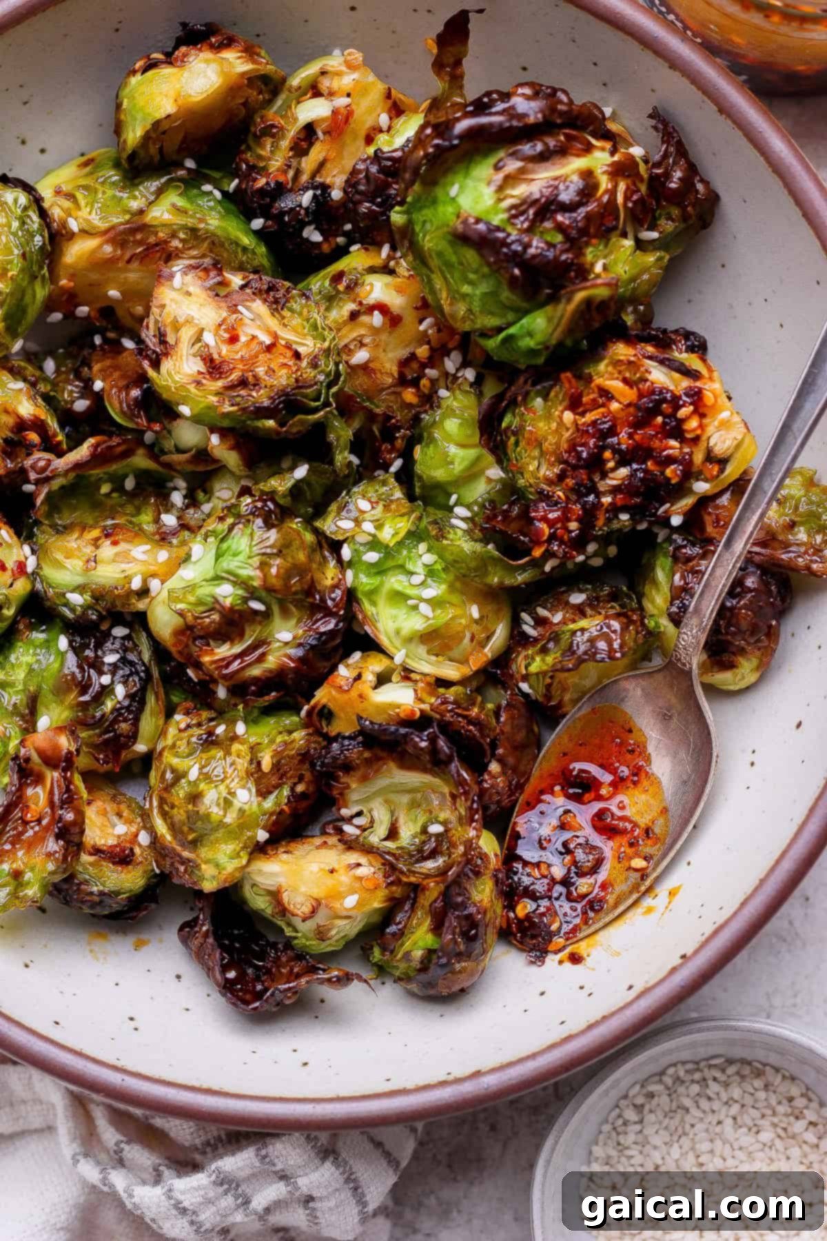 A bowl of roasted Brussels sprouts garnished with sesame seeds, served with a spoon and chili oil.