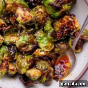 Roasted Brussels sprouts with a glossy chili sauce, garnished with sesame seeds, served in a bowl with a spoon.
