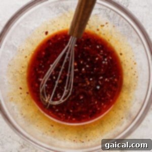 A glass bowl containing a reddish-brown liquid with visible spices and a metal whisk with a wooden handle resting inside.