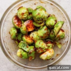 A glass bowl filled with halved Brussels sprouts coated in a dark seasoning or marinade, placed on a light textured surface.