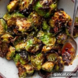 A bowl of roasted Brussels sprouts topped with sesame seeds and chili oil, with a spoon on the side.
