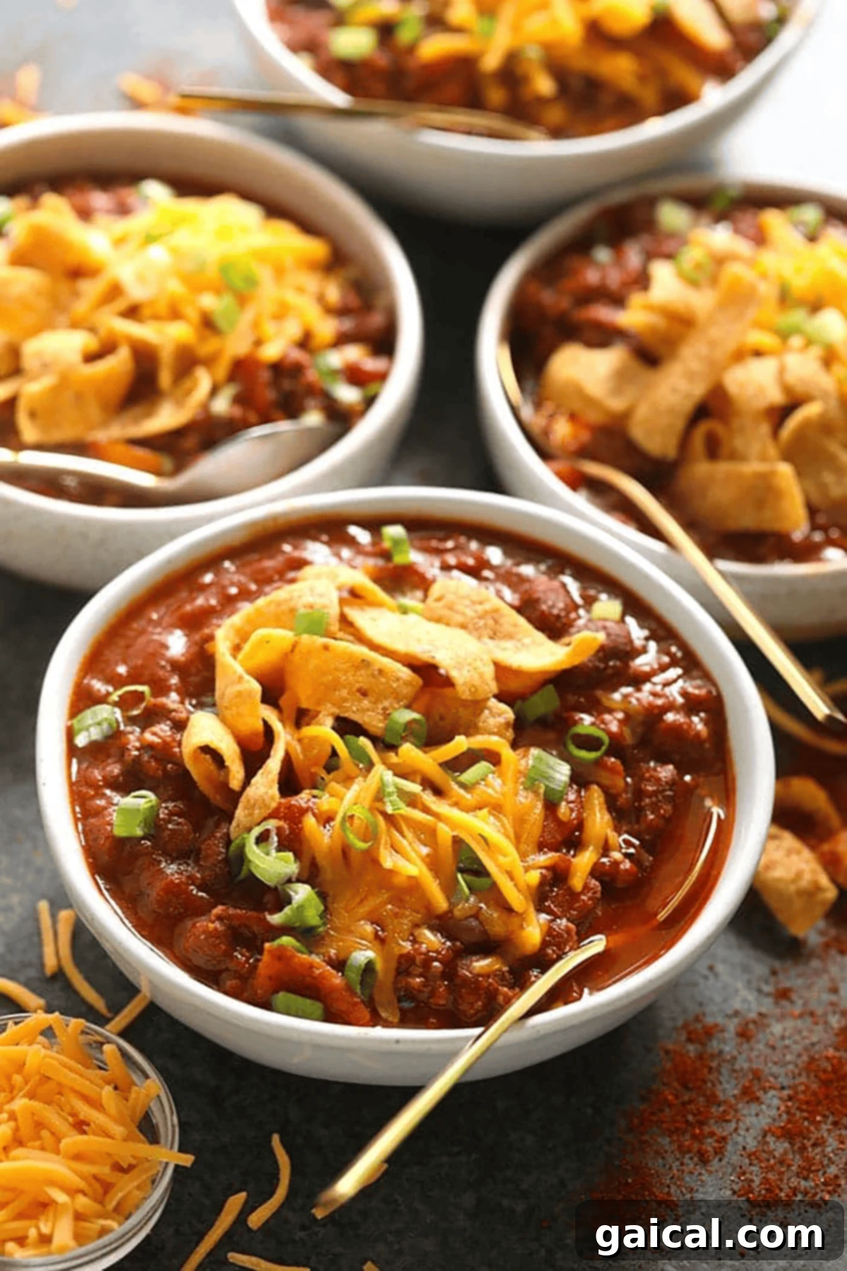 Çok Ödüllü Şampiyon Chili 2 Four bowls of chili topped with shredded cheddar cheese, corn chips, and chopped green onions, with a side of shredded cheese and gold spoons. A perfect chili recipe for any occasion.