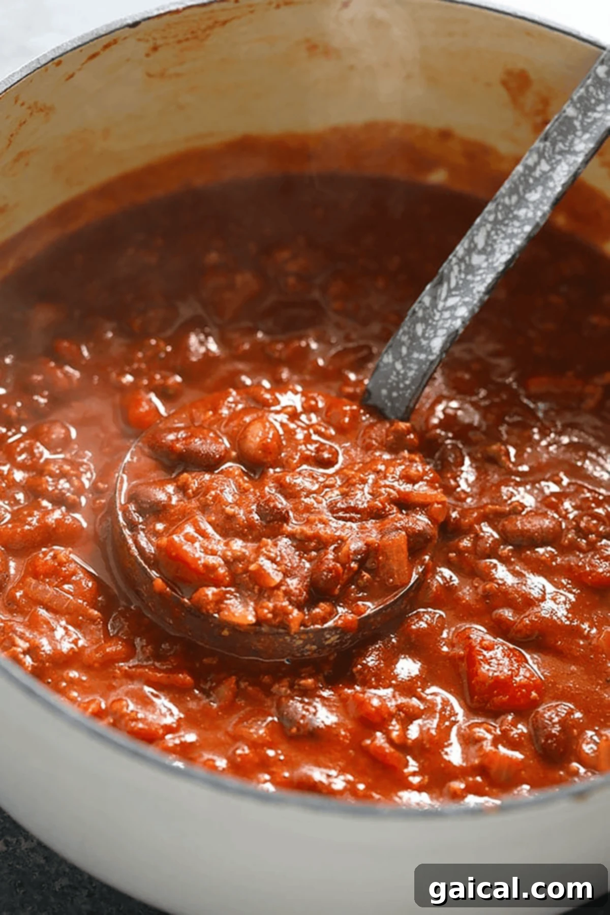 Çok Ödüllü Şampiyon Chili 3 A ladle scoops thick chili with beans, tomato chunks, and ground meat from a large white pot—an inviting chili recipe in progress.
