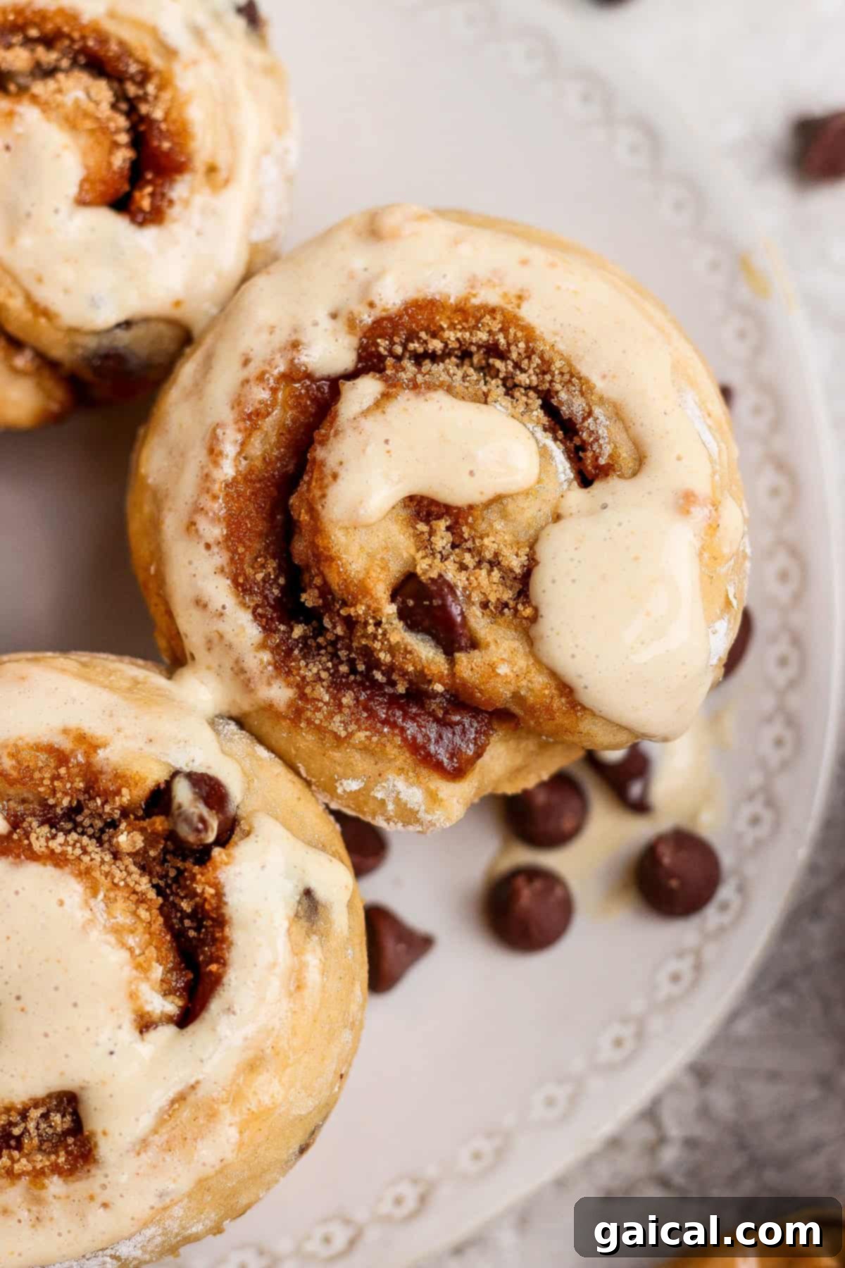 Close-up of freshly baked cinnamon rolls topped with sweet icing on a decorative white plate, with chocolate chips scattered nearby, showcasing an irresistible high-protein dessert.