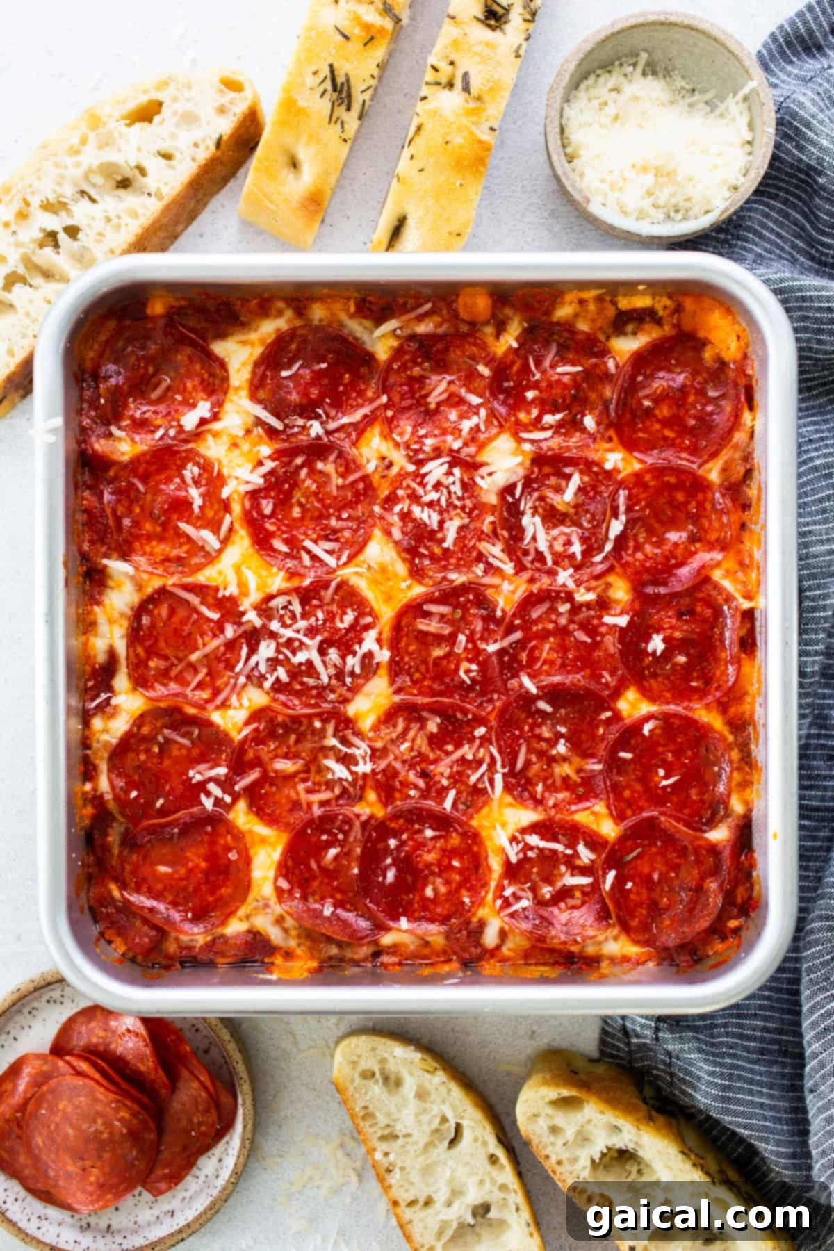 A square deep-dish pepperoni pizza baked in a pan, surrounded by crusty bread slices, a bowl of grated cheese, and a small plate of pepperoni slices on a light surface, inviting a group to share a delectable cottage cheese pizza dip.
