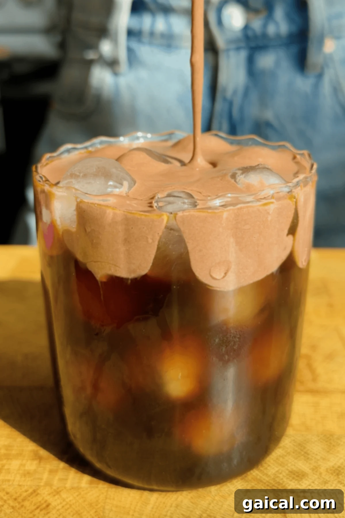 Starbucks Protein Boost Cold Foam 2 A glass of iced coffee topped with whipped chocolate cream, with someone pouring more cream on top. Denim clothing is visible in the background.