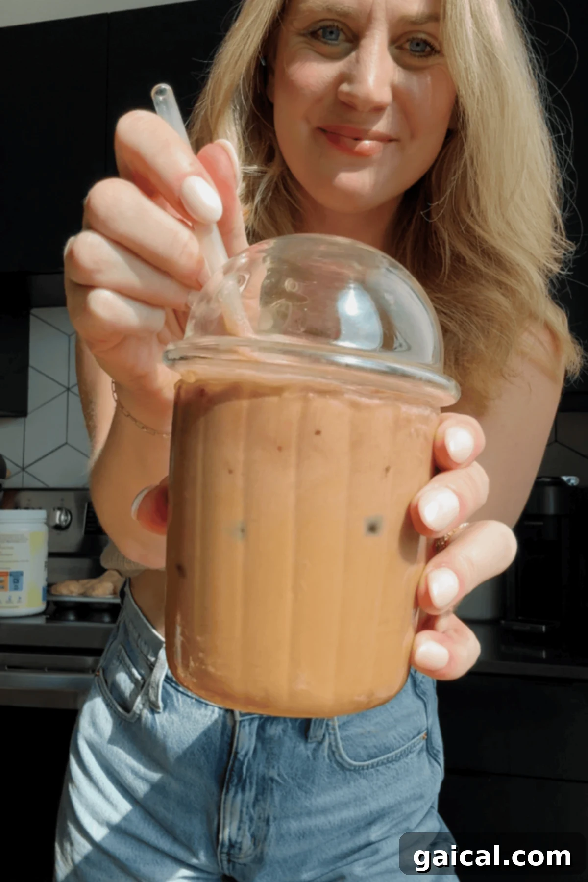 Starbucks Protein Boost Cold Foam 3 A woman holds a plastic cup of iced coffee with a dome lid and straw, topped with protein cold foam, standing in a kitchen.