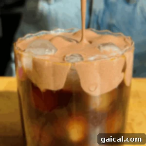 Starbucks Protein Boost Cold Foam 4 A glass of iced coffee topped with whipped chocolate foam, with the foam being poured over the ice. Denim fabric is visible in the background.