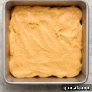 A square metal baking pan filled with an even layer of light brown cake batter on a textured light surface.