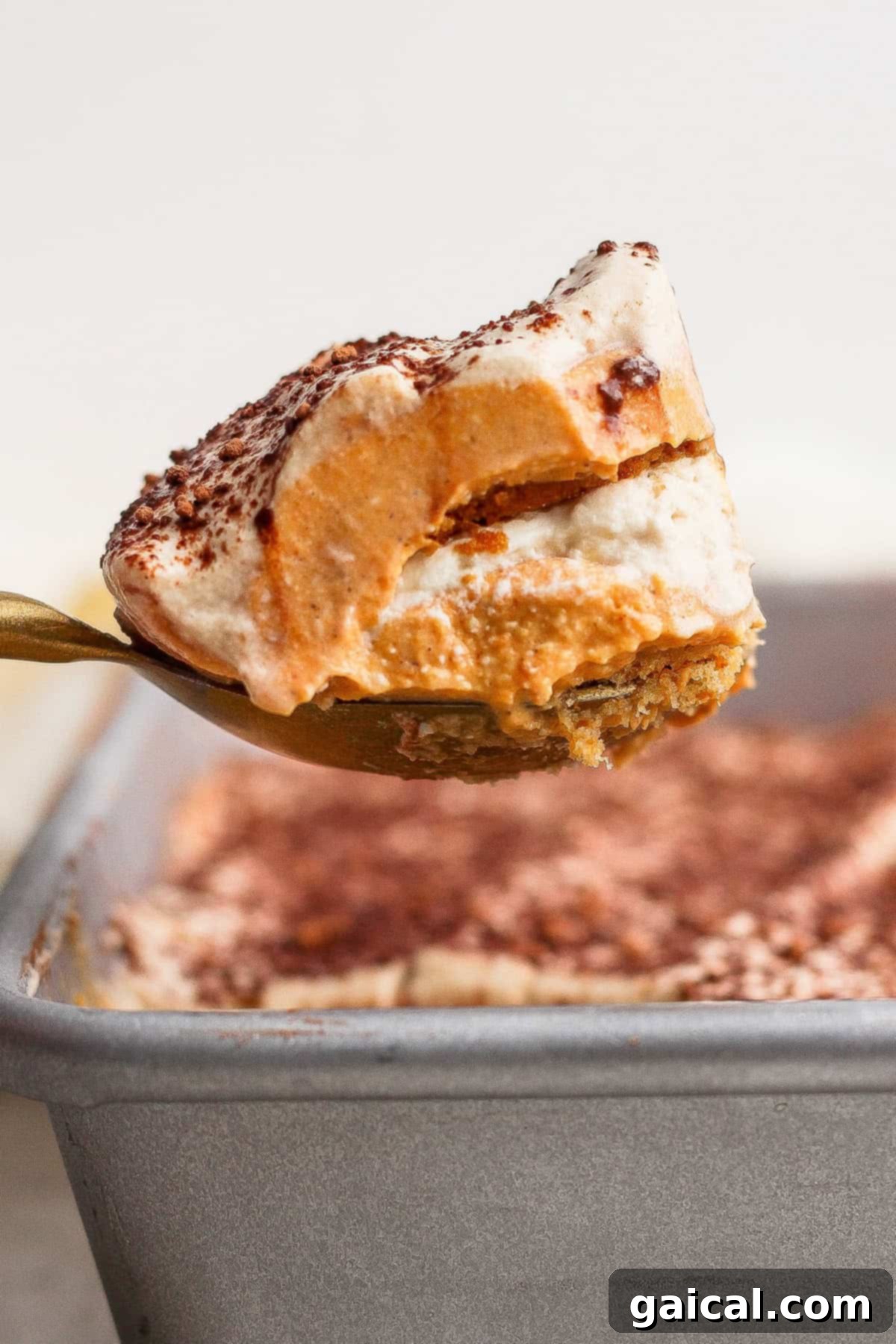 Protein-Powered Pumpkin Tiramisu Autumn Delight 4 A spoon lifts a layered dessert with a creamy top dusted with cocoa powder from a baking pan.