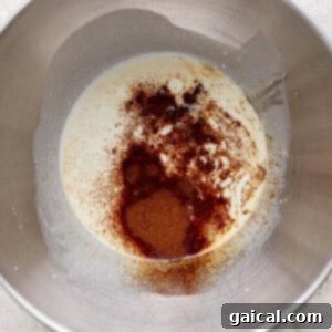 A metal mixing bowl contains a mixture of cream and sprinkled ground spices, including cinnamon, on a light textured surface.