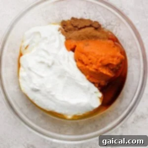 A glass bowl containing pumpkin puree, Greek yogurt, maple syrup, and spices on a light textured surface.