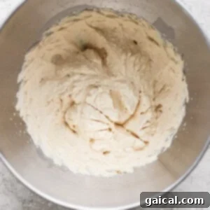 A metal mixing bowl containing light-colored creamed mixture, likely butter and sugar, on a textured gray surface.