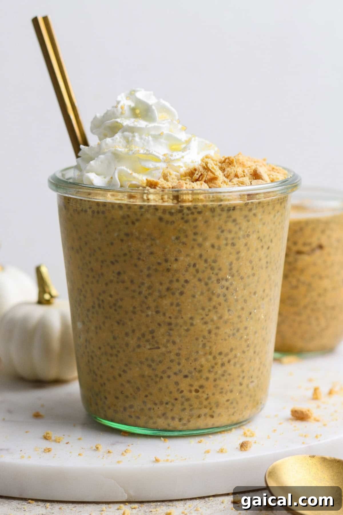 A glass filled with pumpkin chia pudding, topped with whipped cream and crushed crumbs, with a gold spoon and straw, on a white surface.