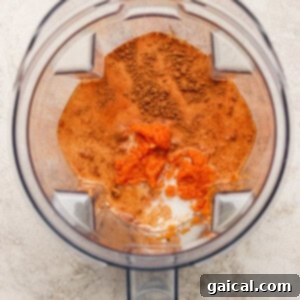 Overhead view of a blender containing orange puree, milk, and brown spices on a light-colored surface.