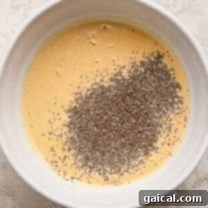 A white bowl containing whisked eggs with a pile of chia seeds on top, set on a light-colored textured surface.