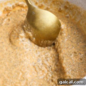 A gold spoon stirs a thick, light brown chia pudding mixture in a white bowl.