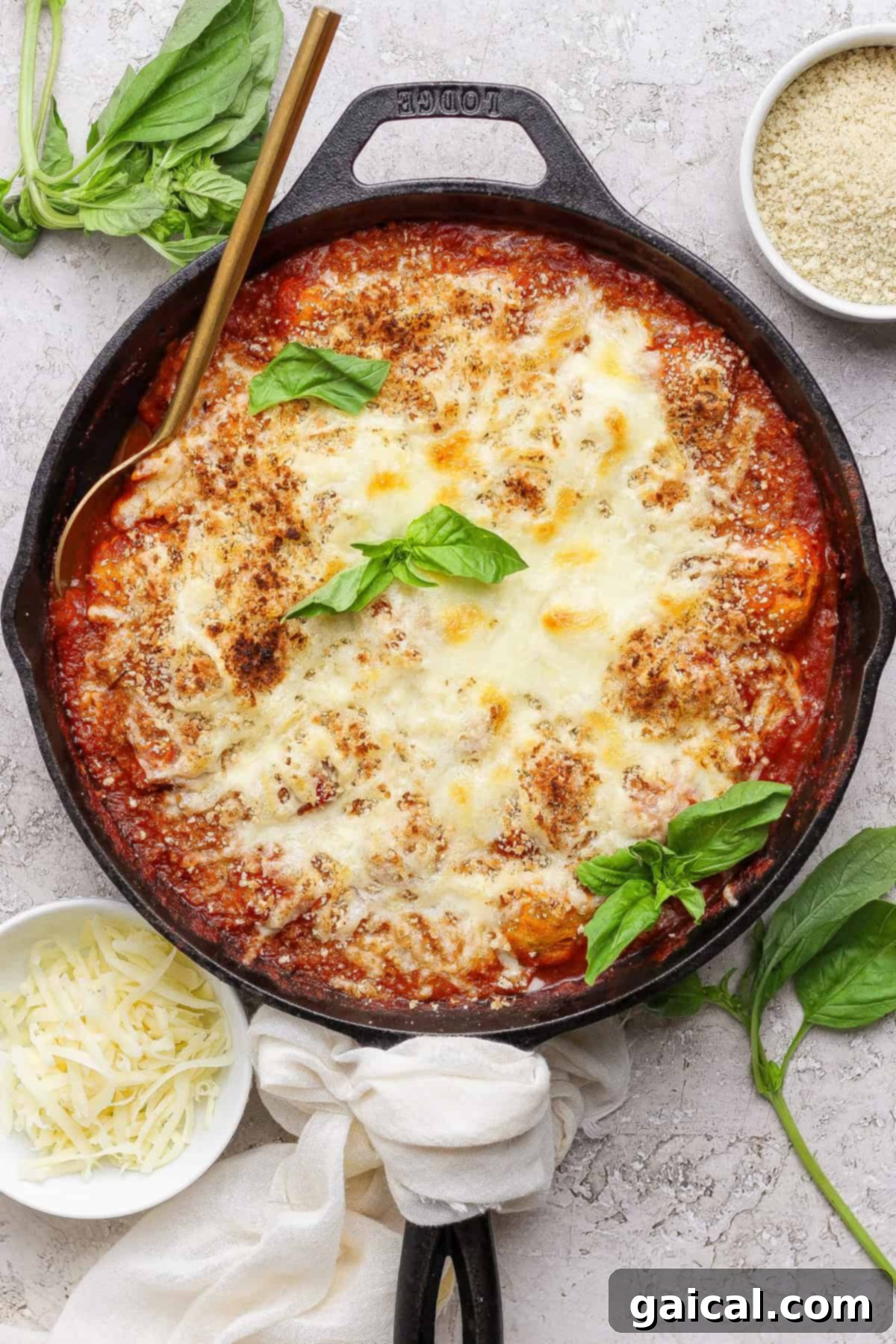 One Pan Parmesan Chicken Meatball Feast 2 A cast iron skillet filled with baked cheesy tomato dish, garnished with fresh basil, surrounded by small bowls of shredded cheese and breadcrumbs.