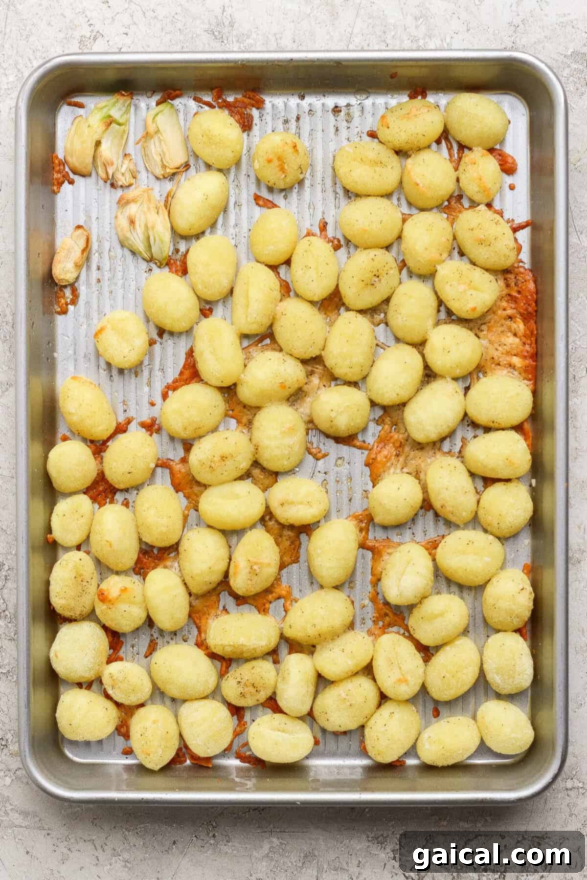Roasted Gnocchi Kale Power Bowl with Garlic Parmesan 3 A metal baking sheet showcasing cooked, golden-brown gnocchi, interspersed with clumps of melted Parmesan cheese and a few roasted garlic cloves on a light, textured surface.