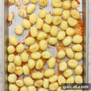 A metal baking sheet with scattered cooked gnocchi, some clumps of melted cheese, and a few roasted garlic cloves on a textured surface, showcasing their golden crispness.