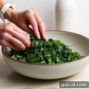 A person massages chopped kale in a large bowl, with a white bottle in the background on a light-colored surface, illustrating the tenderizing process.