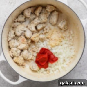 A white pot containing cooked chicken, chopped onions, minced garlic, and tomato paste.