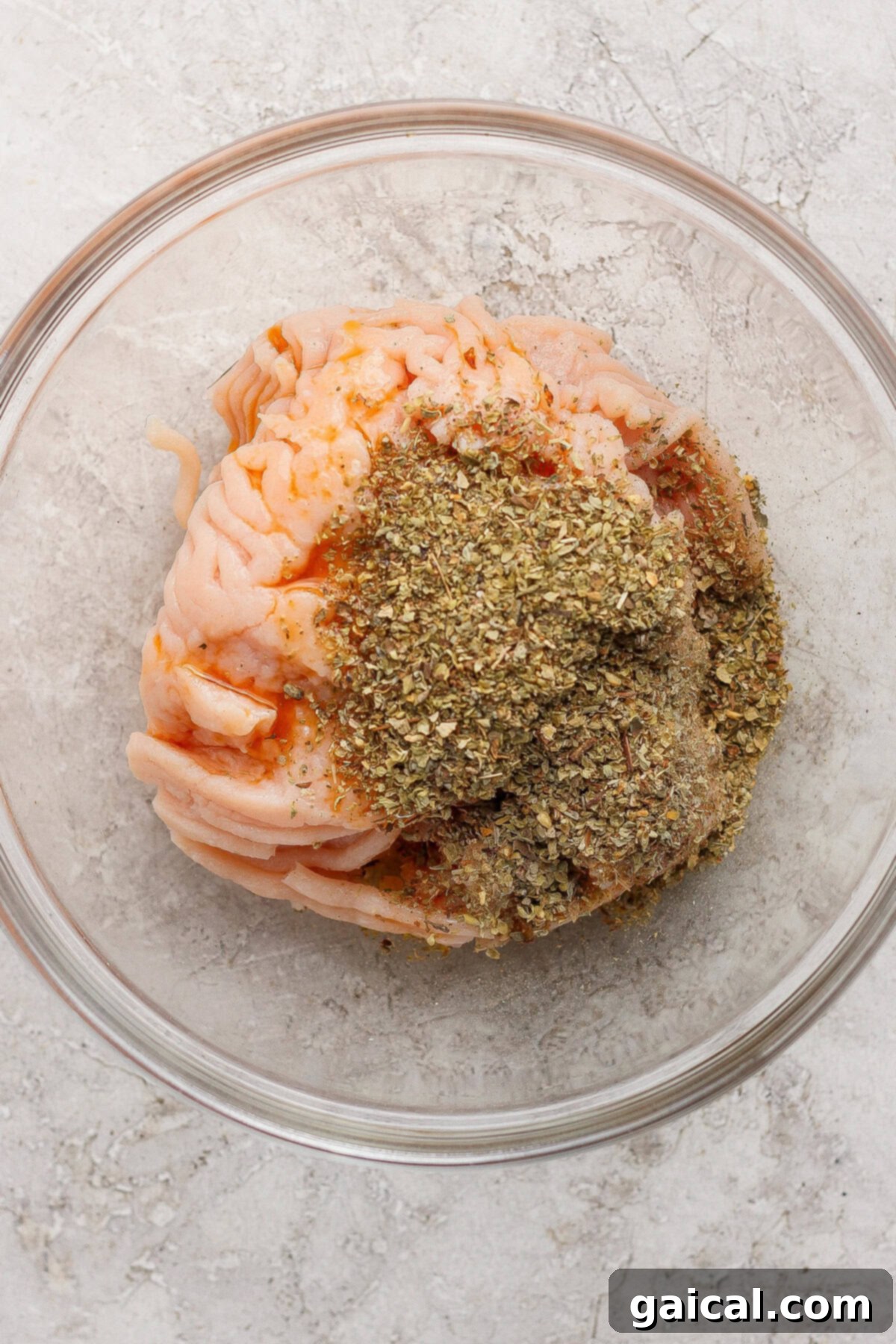 Raw ground chicken seasoned with Italian herbs in a clear bowl, ready for cooking.